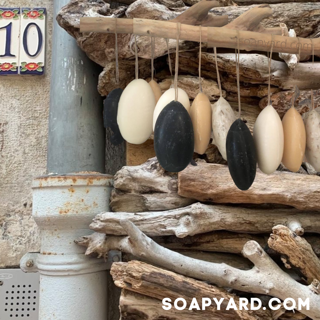  Marseille-style soap bars in natural shades hanging by rope on driftwood, photographed in a rustic French setting with the number "10" in the background.