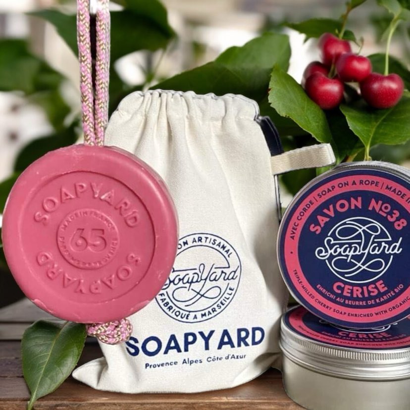 "Cherry Soap on a Rope with matching tin and reusable cotton bag – a stylish, eco-friendly gift perfect for those who love fruity fragrances." the image has an ai background with some leaves and cherries