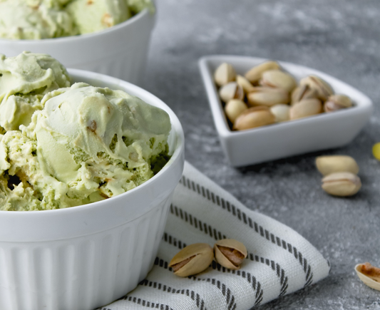 A small white bowl of pistachio ice cream on a white tea towel with blue stripes and a another smaller bowl of pistachios and some pistachios loose on the table. the ice cream is in focus the rest is a little blurred